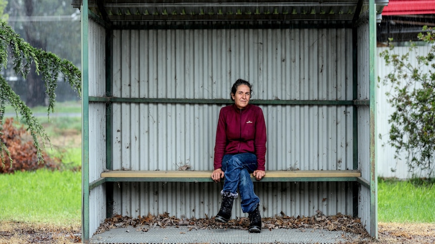 Woman in red jumper and blue jeans with greying black hair sits in tin bush shelter with rain falling