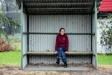 Woman in red jumper and blue jeans with greying black hair sits in tin bush shelter with rain falling