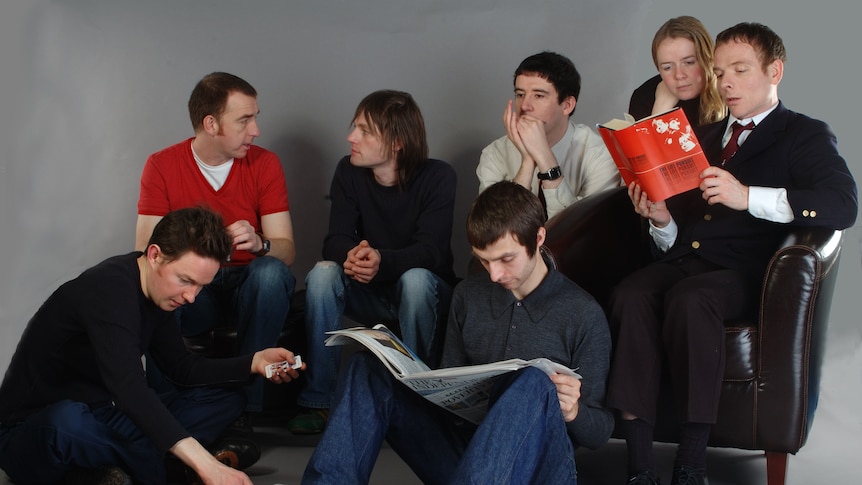 Seven members of Scottish band Belle & Sebastian sit on a chair and the ground reading a book, a newspaper, and playing cards.