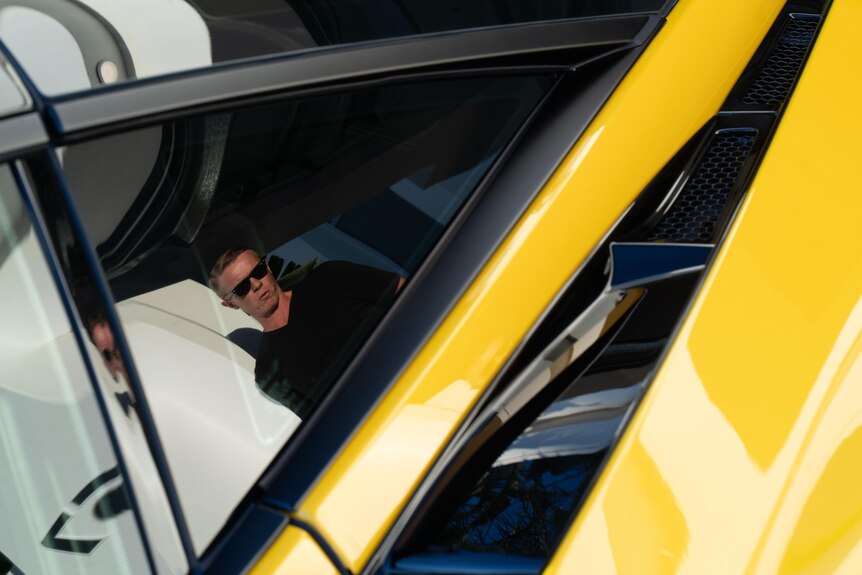 A man in sunglasses is reflected in the tinted windows of an orange car.