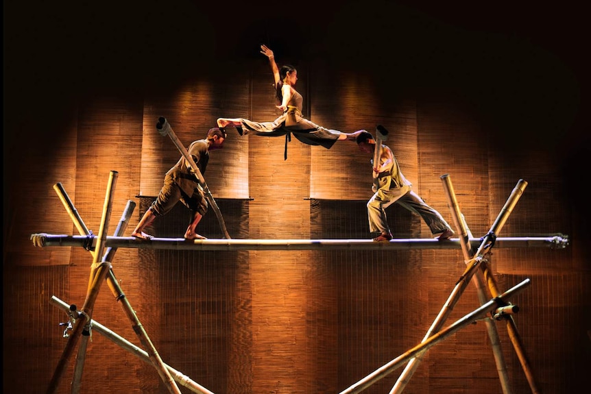 Woman with legs spread balancing a foot each on the heads of two men, all three standing on bamboo structure.