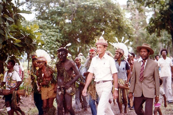 A group of men walk outside together, variously dressed in Papua New Guinean headdresses, suits, and ceremonial wear.