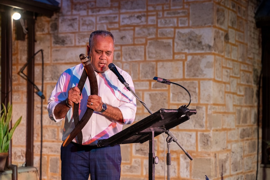 Outdoor evening shot featuring a middle-aged man standing at mic singing and holding two clapping sticks.