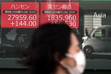 A woman with short dark hair wearing a white face mask walks past a wall of red and green squares with numbers on them.