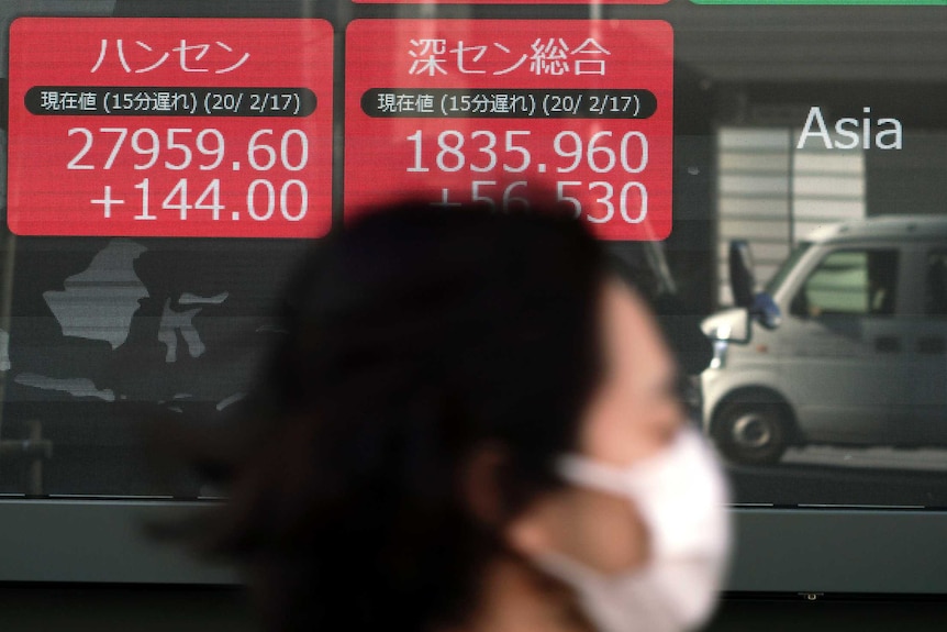 A woman with short dark hair wearing a white face mask walks past a wall of red and green squares with numbers on them.