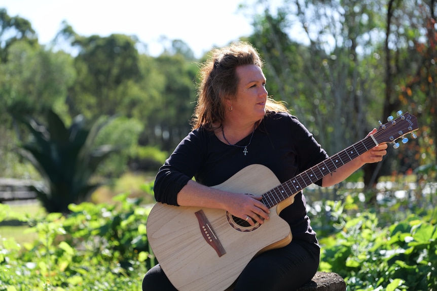 Middle-aged woman in a long black-sleeved shirt sitting in a park and holding a guitar