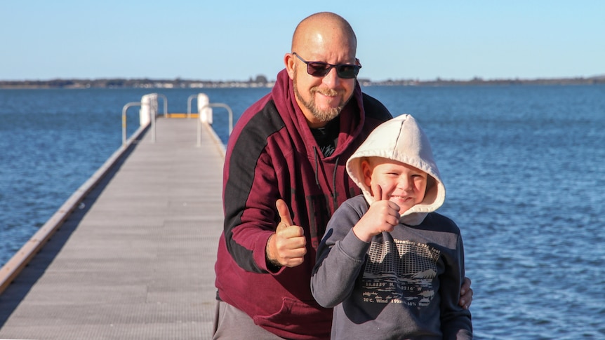 Bald older man,  beard, wears red, black hoodie, smiling boy, white hood up, stand on a jetty, gives thumbs up. Sunny day.