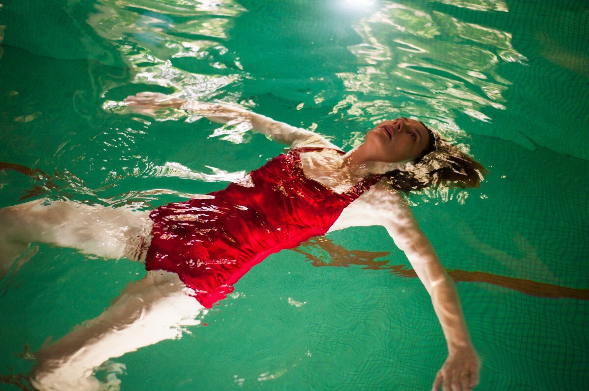 Mid-shot of performer Tiffany Lyndall Knight floating face up in an indoor pool wearing a red swimsuit.