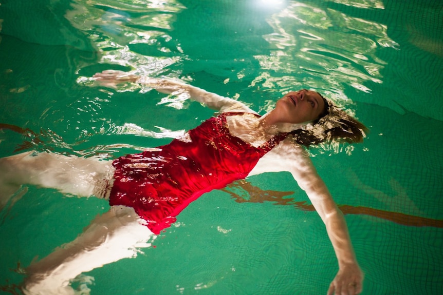 Mid-shot of performer Tiffany Lyndall Knight floating face up in an indoor pool wearing a red swimsuit.