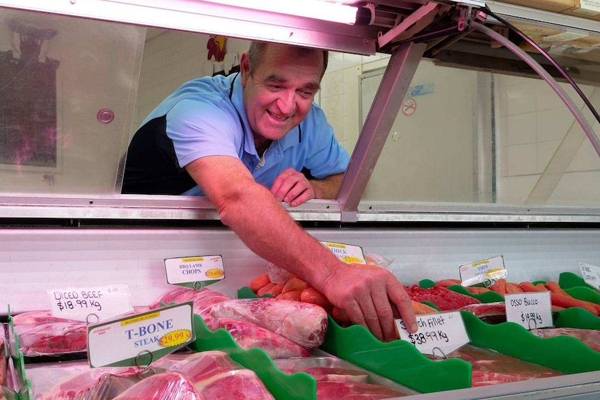 Man with blue polo shirt reaches forward from behind a counter to grab a scotch fillet on display