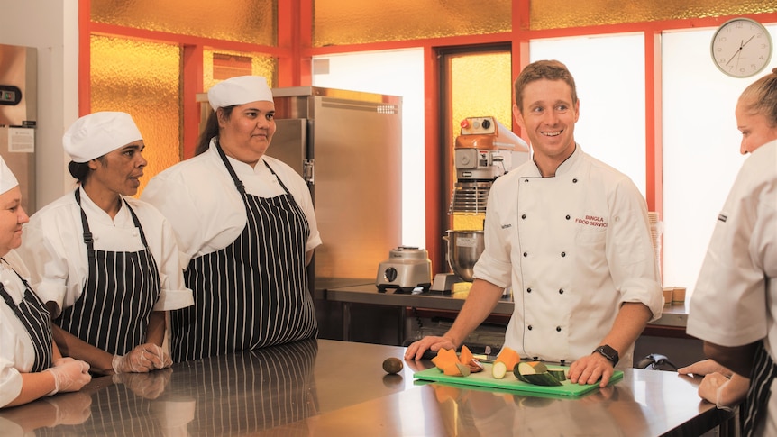 A head chef with trainees in a kitchen learning how to cook.