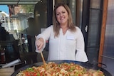 A woman is standing up stirring a large paella pan with a wooden spoon