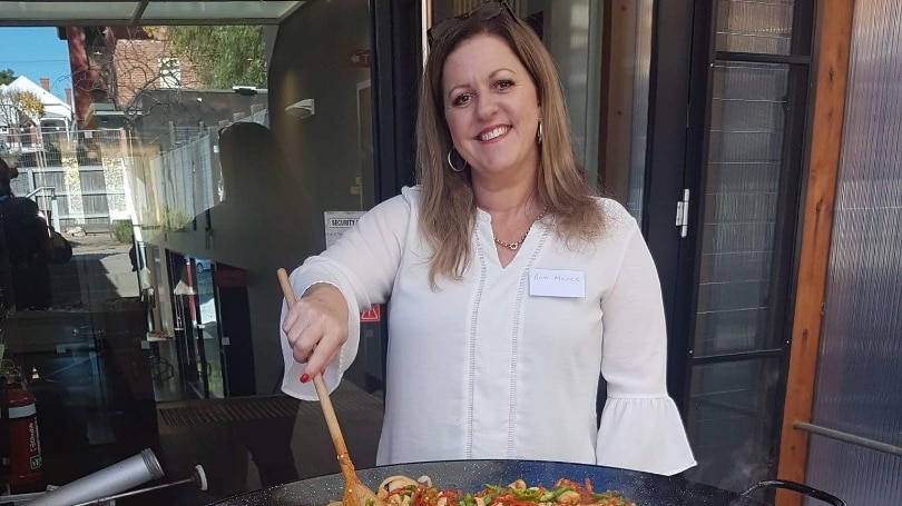 A woman is standing up stirring a large paella pan with a wooden spoon