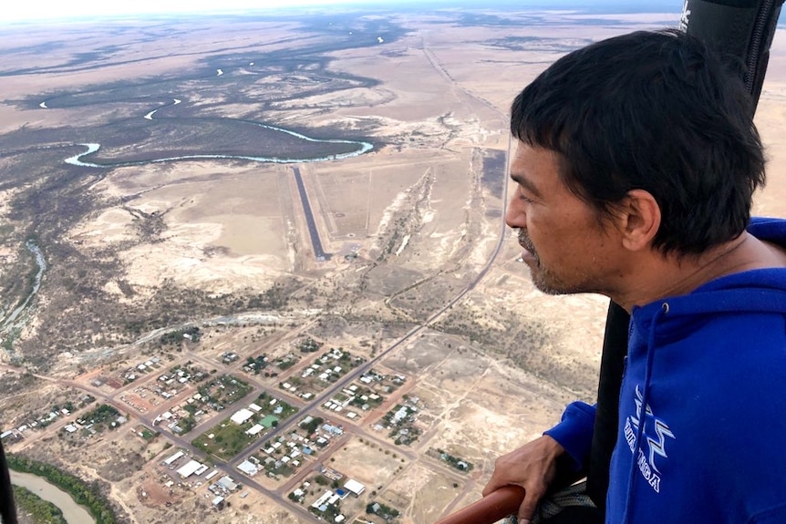 An Indigenous man, wearing a blue shirt, looks out from a hot air balloon onto a township and saltpans