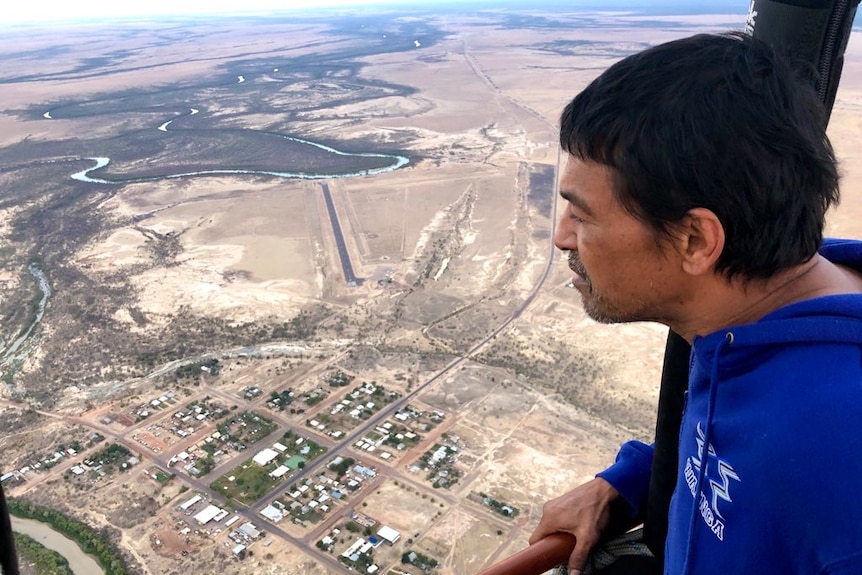 An Indigenous man, wearing a blue shirt looks out from a hot air balloon onto a township and saltpans