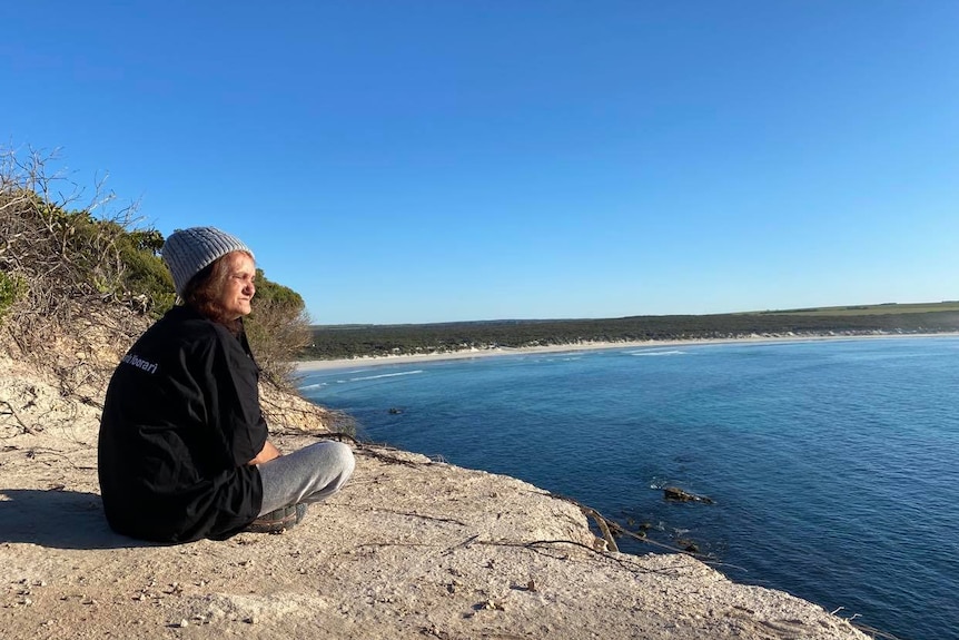 A woman wearing a dark jumper, grey pants and a beanie sits crossed legged looking out over the water