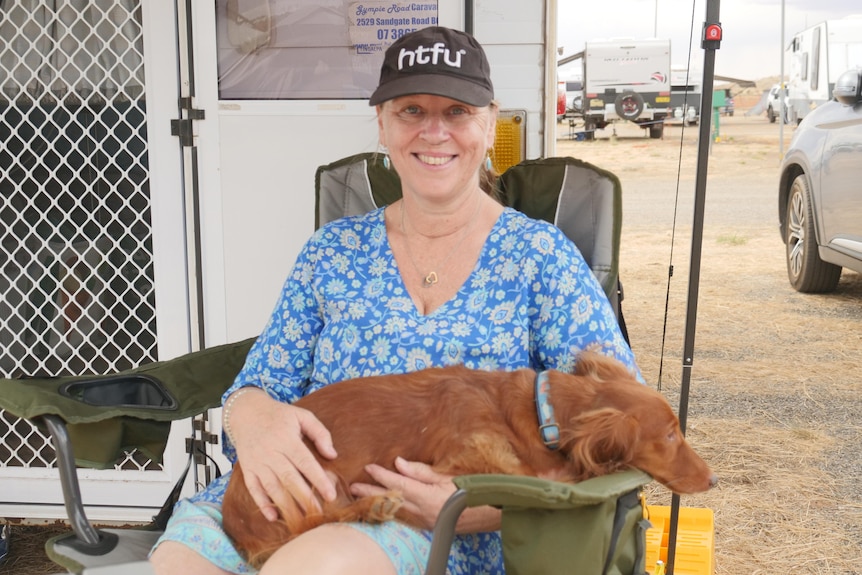 A woman in a blue blouse and black cap sits in a chair with a small red dog.