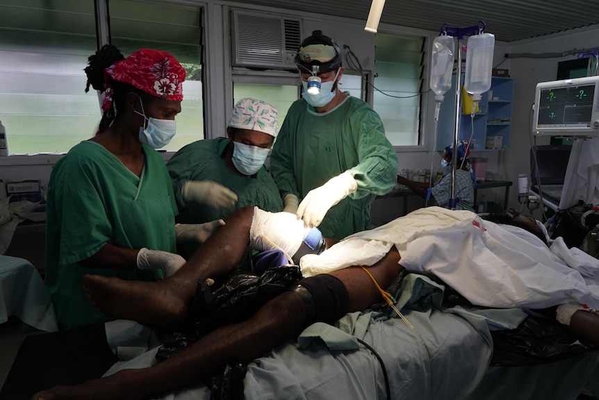 Three health workers wearing surgical gowns work on a patient's upper thigh