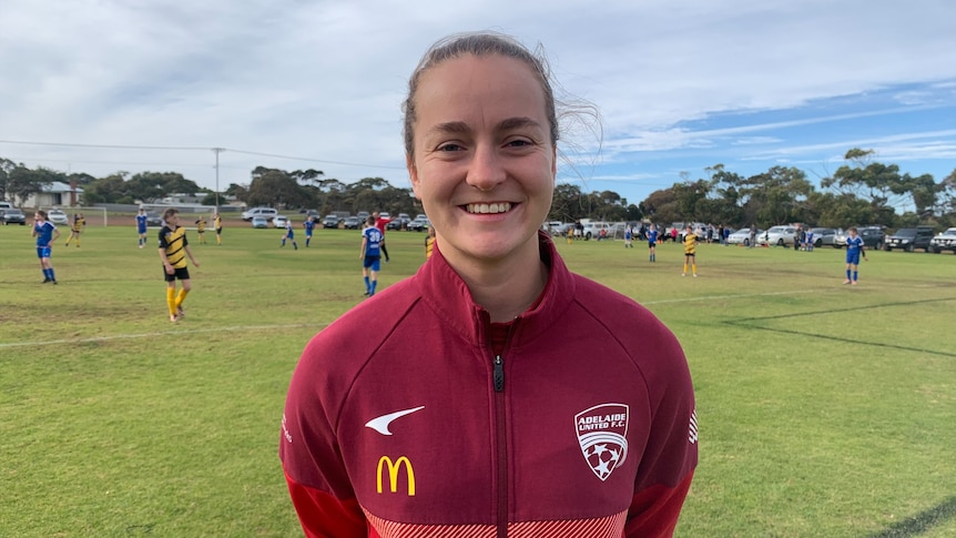 Adelaide United Women’s FC Captain, Isabel Hodgson smiling in a red jumped in front of soccer field.