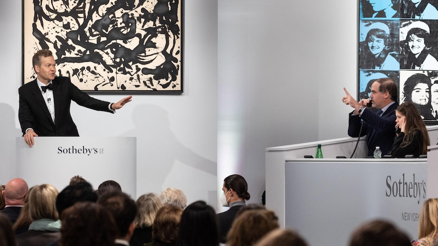 A man stands in front of a Jackson Pollock painting and gestures at another man bidding at an auction.
