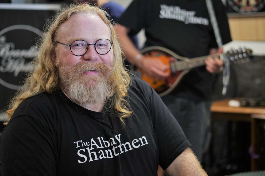A bearded, bespectacled man with long ginger locks wearing a black shirt that says "The Albany Shantymen".