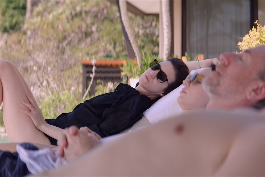 White woman with dark hair and sunglasses wears black sun shirt and reclines on deck chair beside two other people