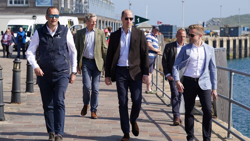 A tall, balding middle-aged white man in glasses and a suit walks on a seaside footpath with a crowd of people.