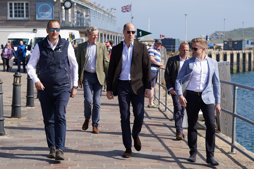 A tall, balding middle-aged white man in glasses and a suit walks on a seaside footpath with a crowd of people.