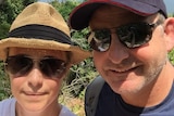 A woman in a beige straw hat and a man in a cap take a selfie with a bay and beach in the background.