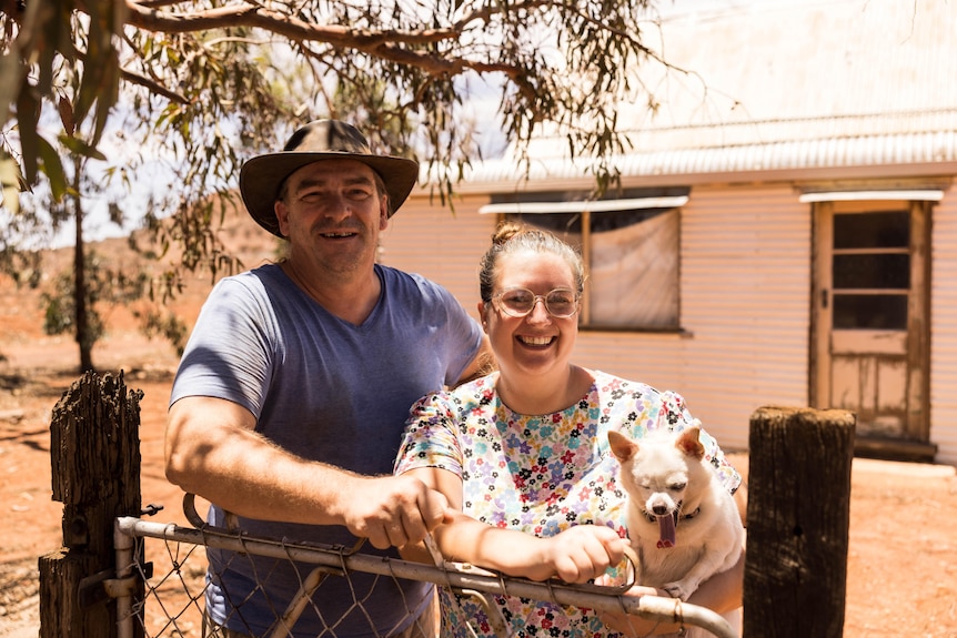 A couple standing at a gate out the front of their outback house.  