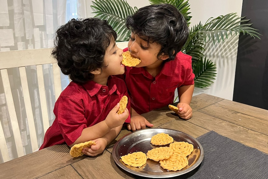 Preethi's sons share a savoury Indian snack, each holding it in their mouth.