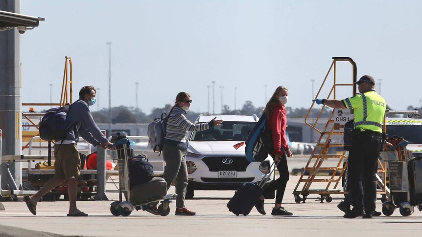 Three passengers wearing face masks and carrying luggage walk towards a police officer and a man on the tarmac at Perth Airport.