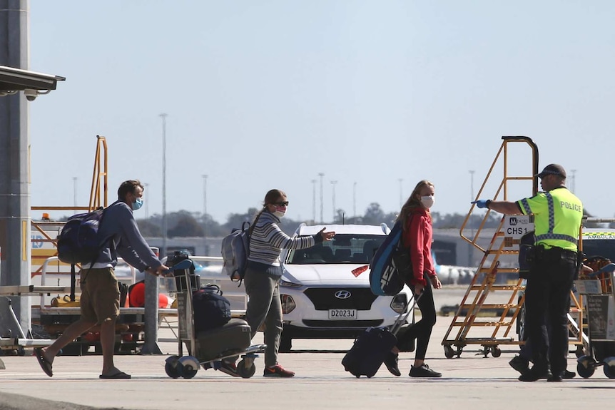 Three passengers wearing face masks and carrying luggage walk towards a police officer and a man on the tarmac at Perth Airport.