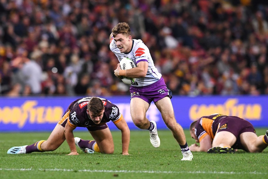 A male NRL player runs with the ball held with bis left arm as two opposition players lie on the ground behind him.