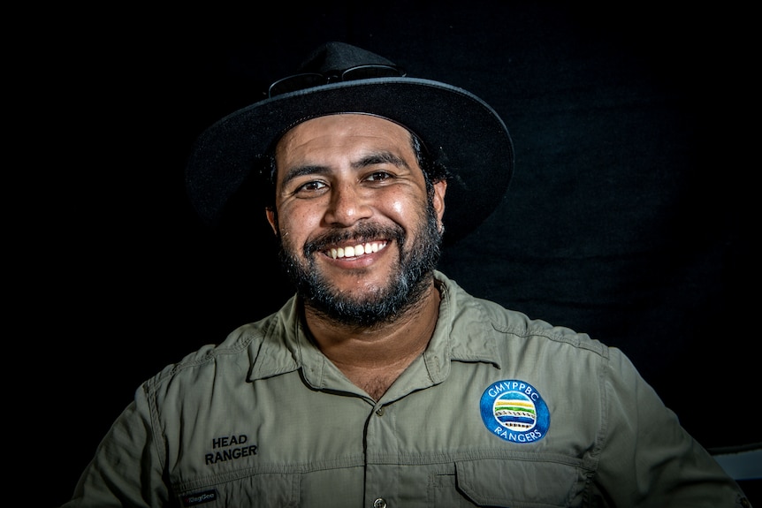 An aboriginal man in a khaki shirt and wide brim hat smiling at the camera.