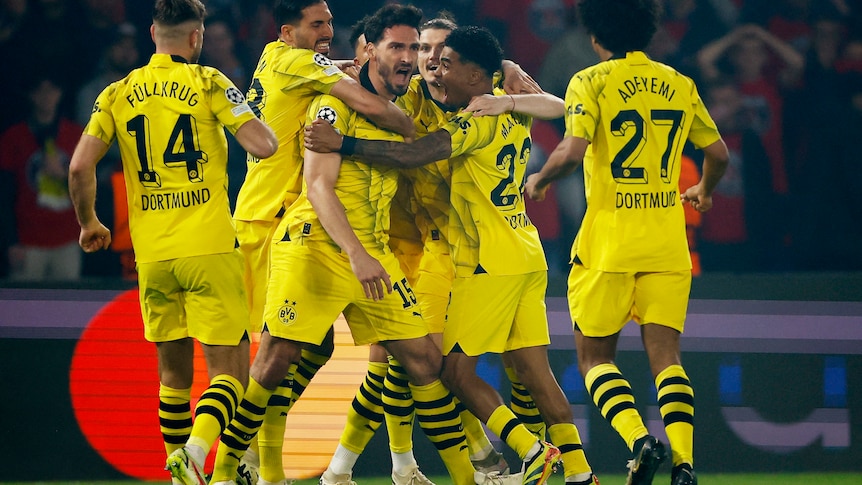 Footballers in yellow jerseys surround a goal scorer, screaming in excitement.