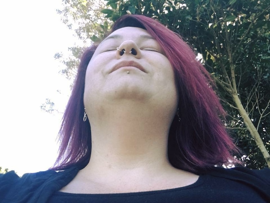 A woman with purple hair and a nose ring tilts her head back with her eyes closed.