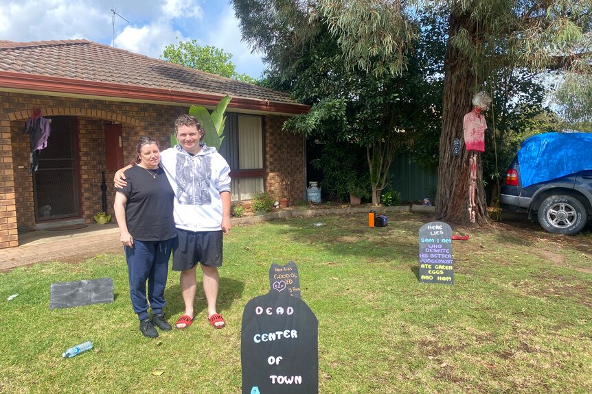 A man and woman standing in front of house with fake gravestones around, one says 'dead center of town'