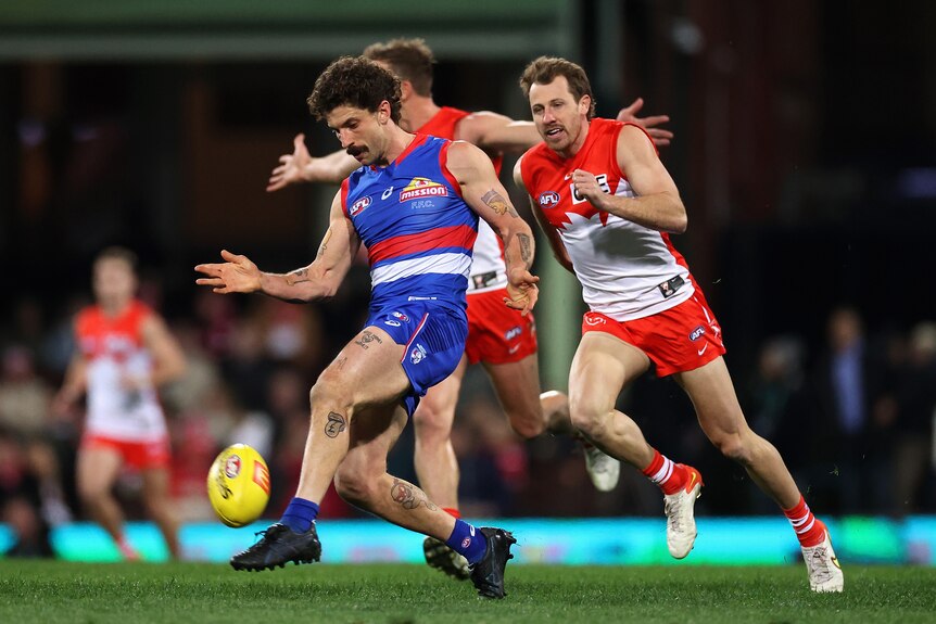 A Western Bulldogs player drops the ball onto his boot to kick it clear, as two Sydney defenders chase behind.