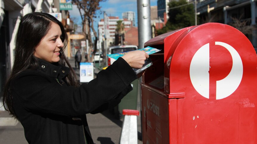 A woman places a colourful envelope in a red Australia Post post box.