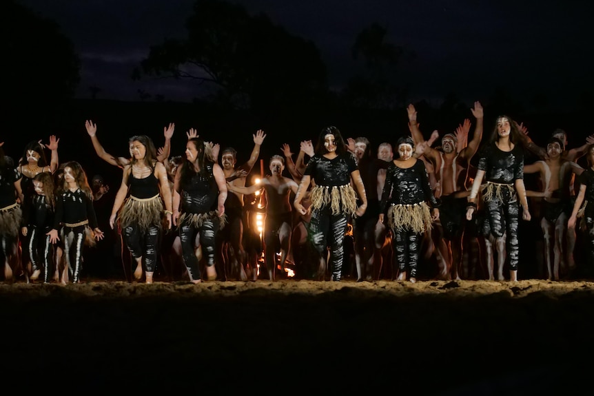 A number of girls and women stand in front on black men on sand doing a traditional dance.