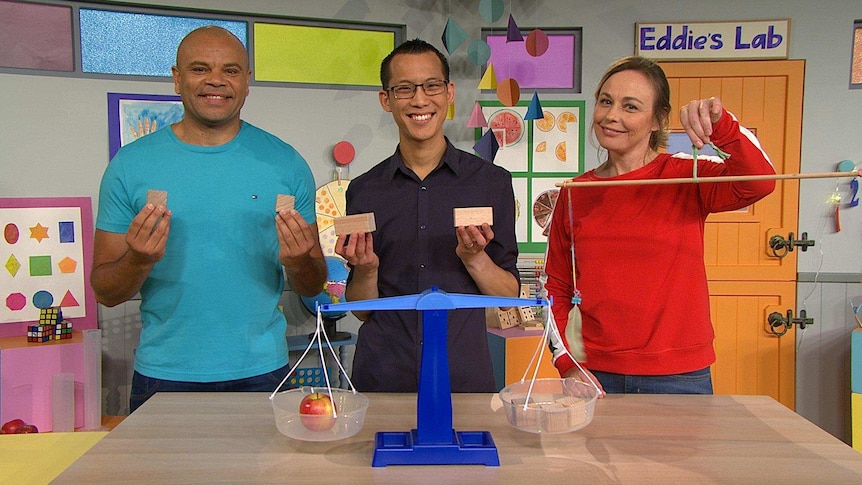 Luke, Abi and Eddie Woo on the Play School set with scales on the desk, holding blocks and a sign "Eddie's Lab" on the wall