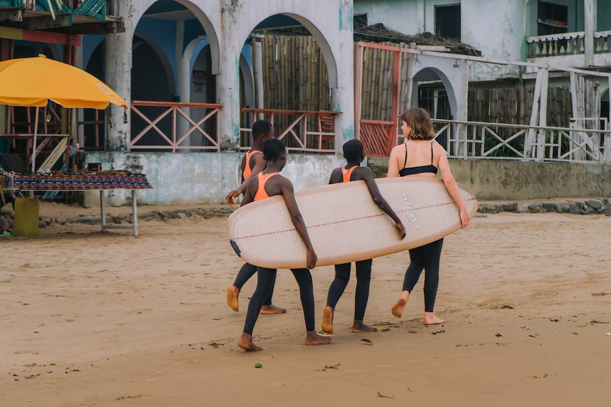 Three women carrying a surf board together on a beach.