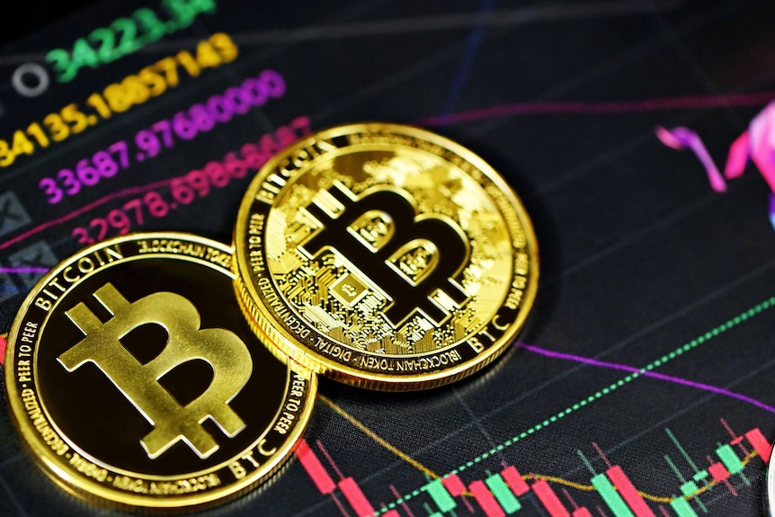 Two large gold coins bearing engraved "b"s lie on top of a complex financial line-and-bar graph