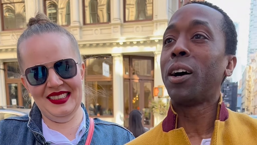 A photo of a woman in sunglasses and red lipstick and a man in a mustard jacket smiling and talking to a camera