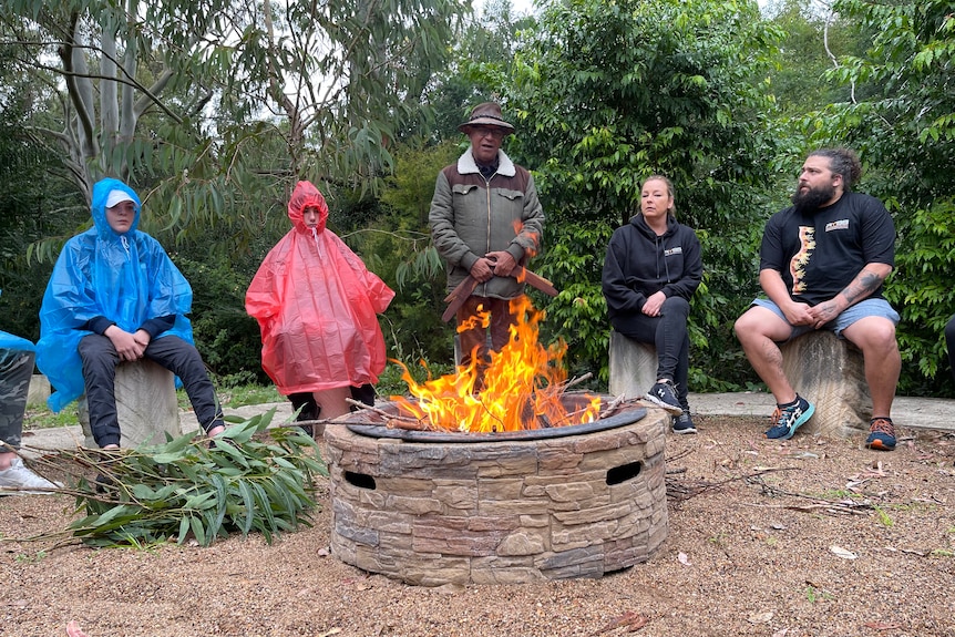 Teenagers in bright raincoats and three adults from the Ngaliya program sit around a fire talking.