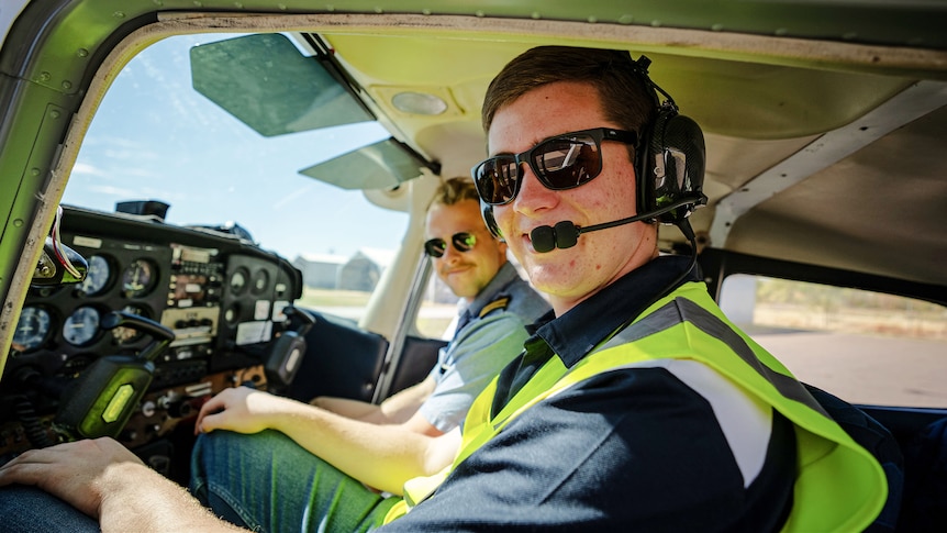 A young man wearing a high-vis vest sits in the small cockpit of a small plane on an airstrip.