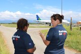 Two women in Australian Aid t-shirt have backs facing camera talking near an approaching Solomon Islands plane.