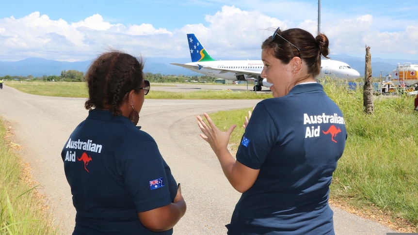 Two women in Australian Aid t-shirt have backs facing camera talking near an approaching Solomon Islands plane.