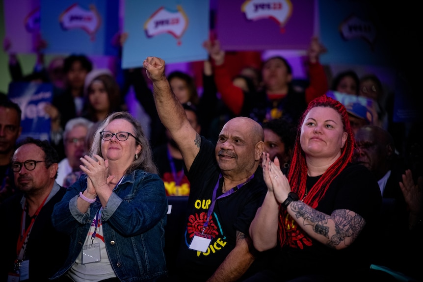 A middle-aged Aboriginal man raises his fist as two women standing beside him clap at a rally for the Yes campaign.